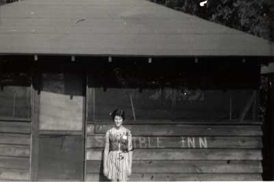 Girl standing in front of cabin at Camp Highland. The words "Stumble Inn" are painted on front of the cabin near the door.